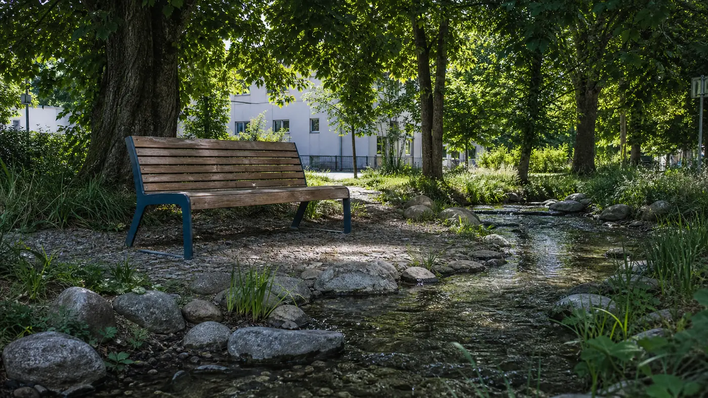Lavička v tieni stromov pri obnovenom potoku v meste